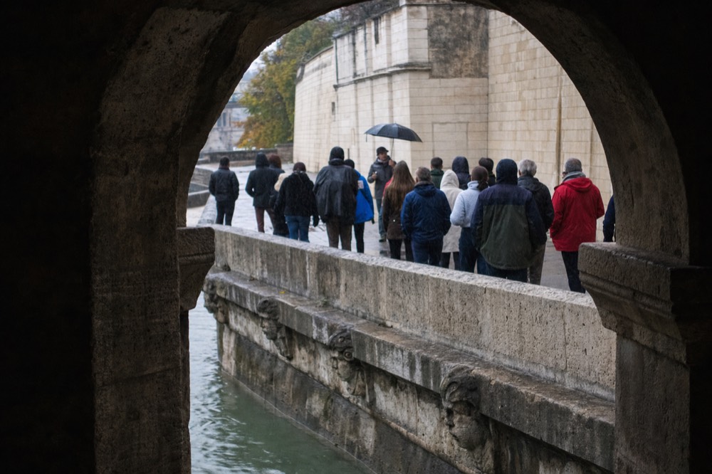 A group listens to a guide with an umbrella beside the Seine, seen through a shadowed stone arch.