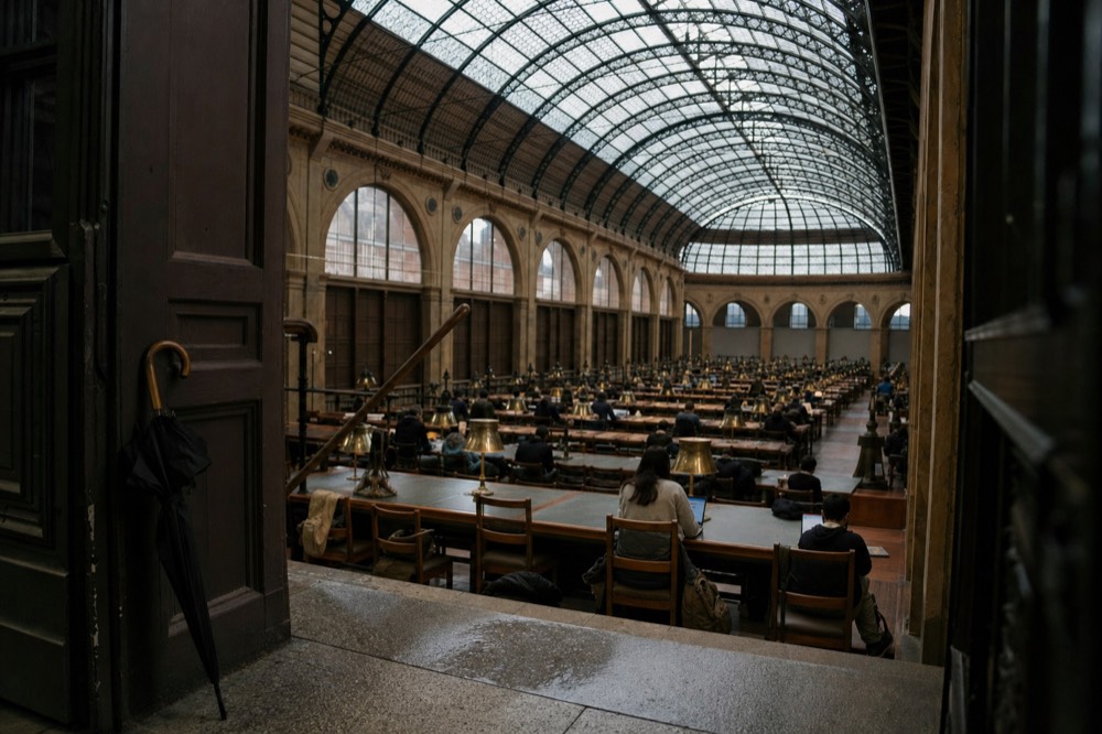 Historic library reading room with a glass-and-iron ceiling, long study desks, brass lamps, seated readers, and an umbrella by the doorway.