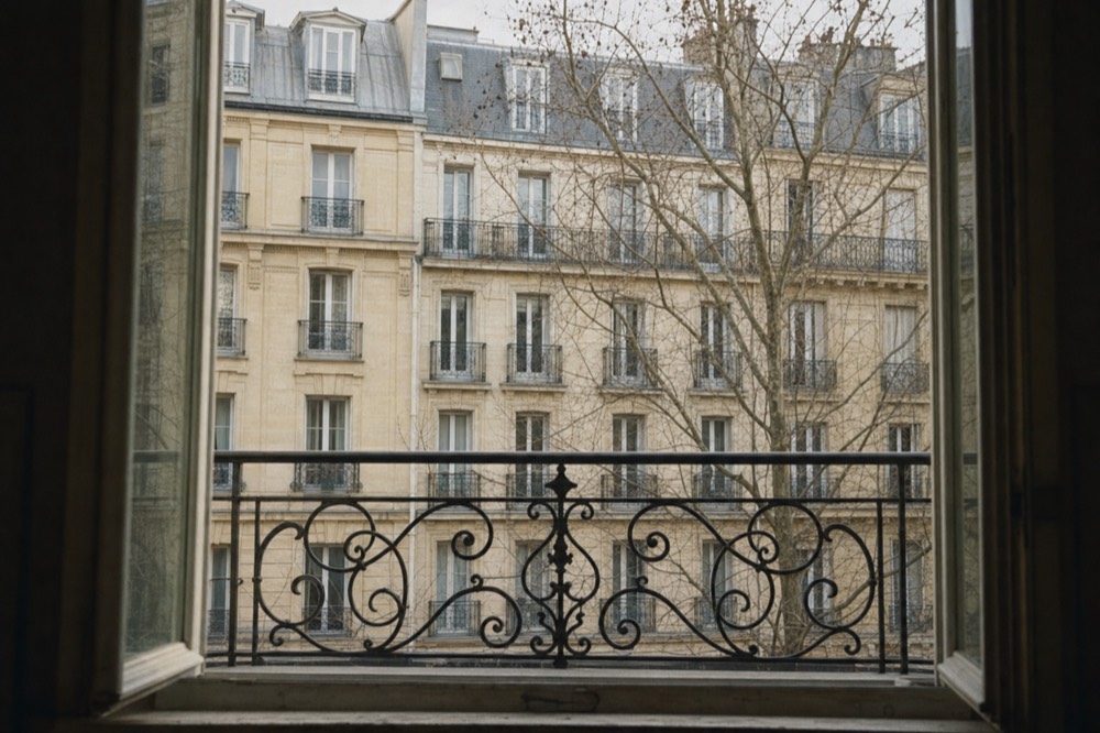Open window with ornate iron railing overlooking cream Paris apartment facades and bare plane tree branches.