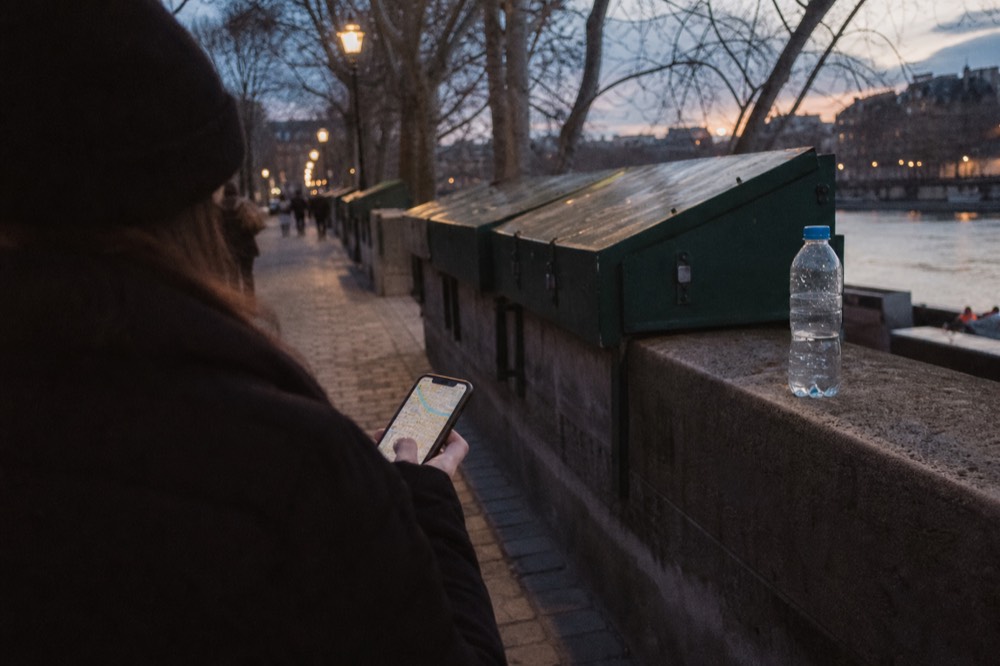 Un participante consulta un mapa en el móvil junto a cajas verdes de buquinistas y una botella de agua en un muelle del Sena al atardecer.