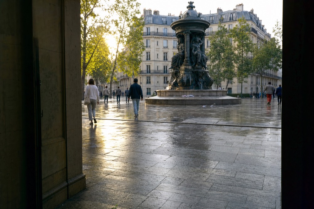 Personas caminan sobre adoquines mojados alrededor de una fuente de bronce oscuro en una plaza de París, enmarcada por un portal en sombra.
