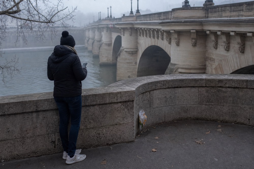 Un paseante solo consulta el móvil junto a un pretil de piedra al lado del Sena brumoso, con arcos de puente pálidos y un envoltorio cerca.