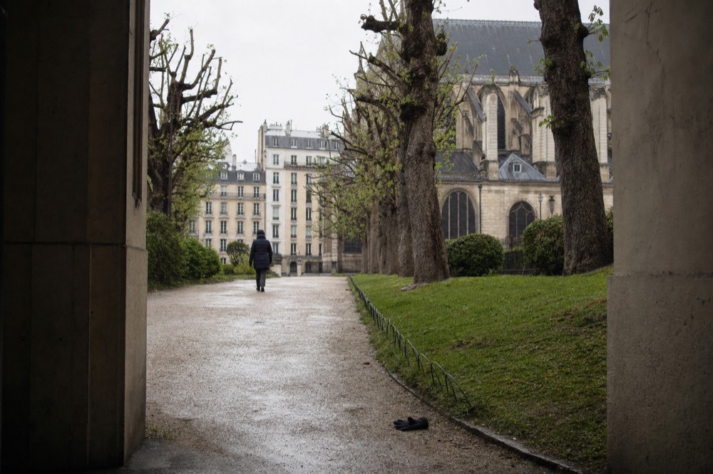 A gravel path with trees leads toward Notre-Dame buttresses, with a lone figure walking away and a dark glove near the grass.
