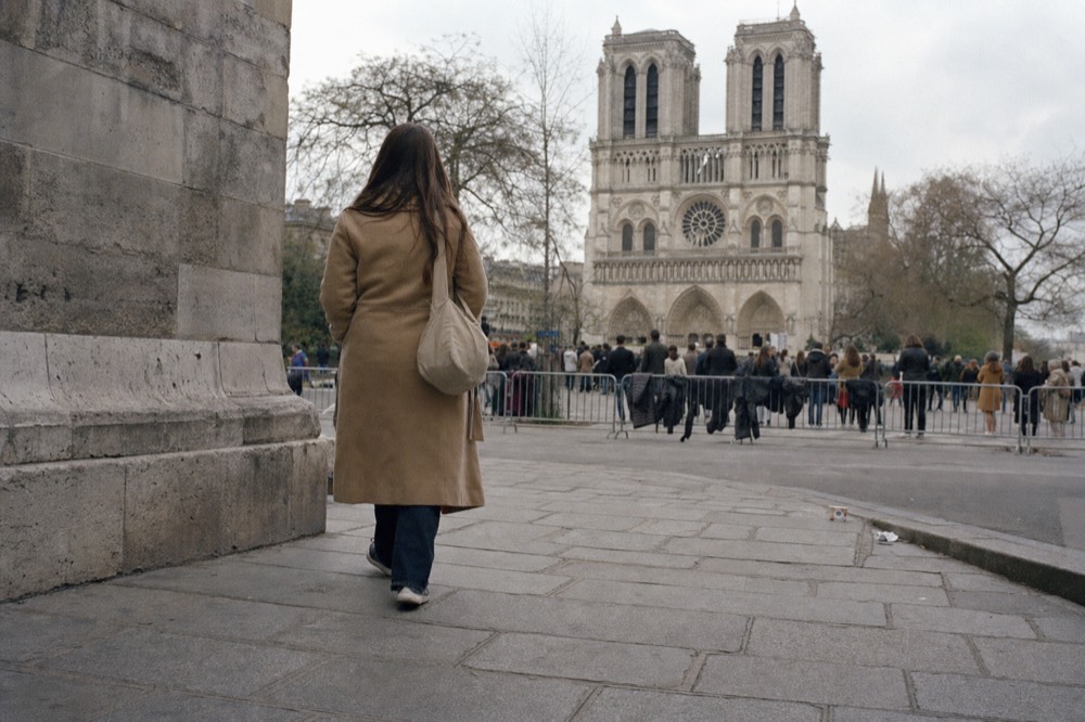 A woman walks on wet paving toward the west front of Notre-Dame, with twin towers, a rose window, crowds, and barriers ahead.