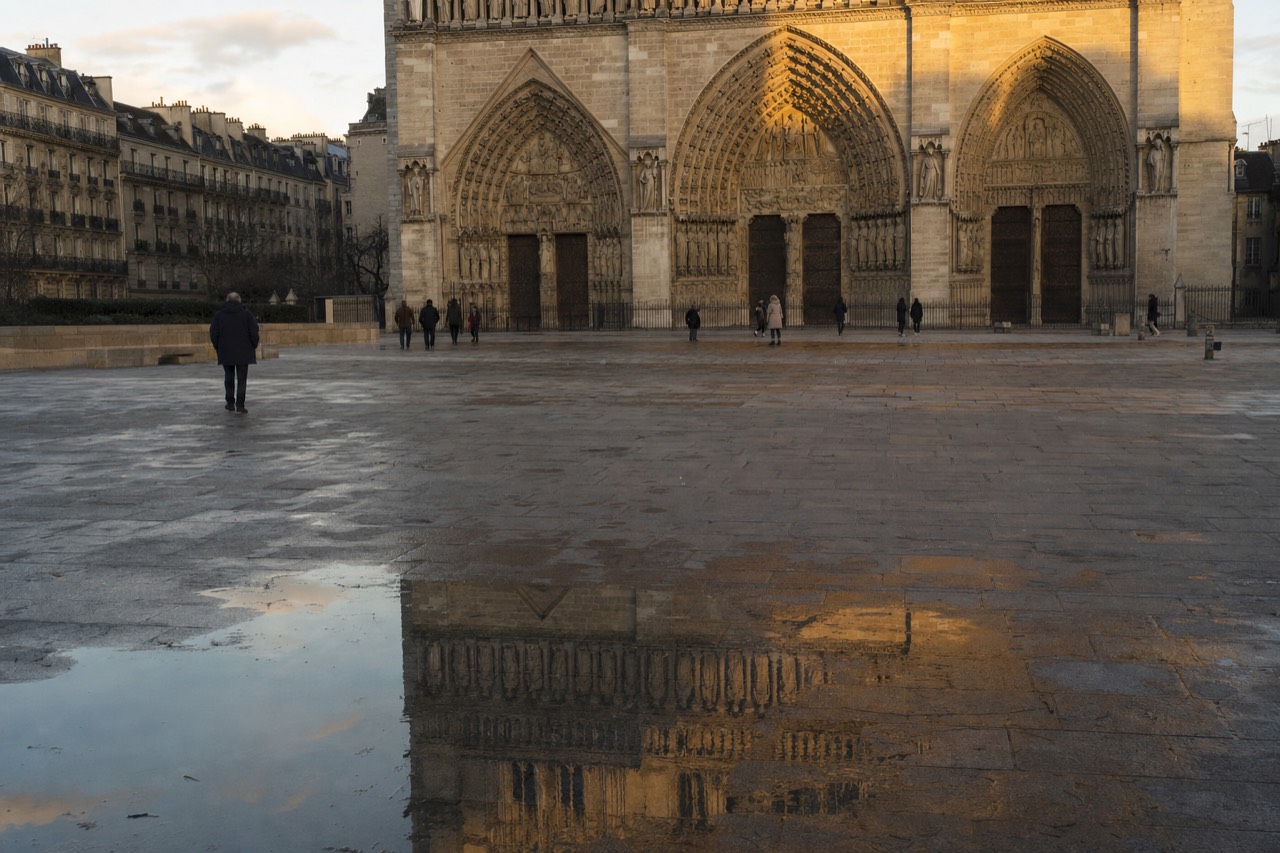 Notre-Dame's western portals face a wet stone parvis with reflections and a few distant pedestrians at golden hour.