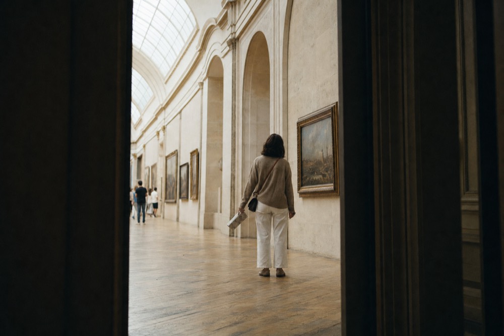 A visitor stands in a vaulted museum gallery lined with framed paintings under a glass roof.