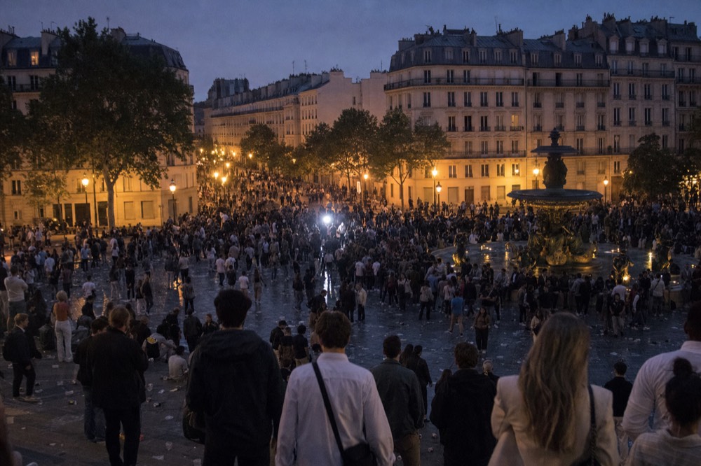 Multitud en una plaza de París al atardecer, alrededor de una fuente, con edificios de piedra caliza y árboles bajo un cielo violeta.