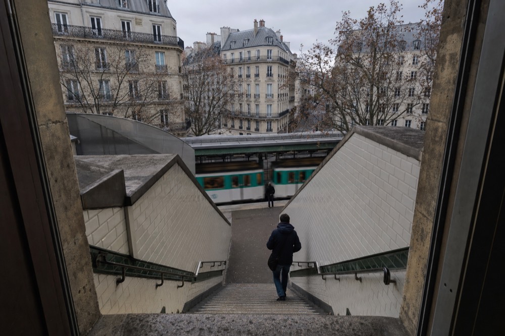 Una escalera de metro con azulejos desciende hacia un andén elevado por el que pasa un tren verde y blanco, con bloques de apartamentos de París al fondo.