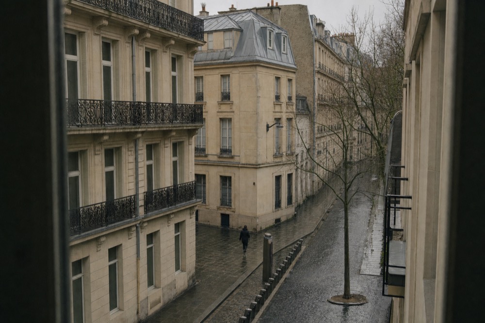 Estaciones de alquiler de bicicletas vacías alineadas en una calle empedrada y mojada de París, bajo fachadas de piedra caliza y balcones de hierro forjado.
