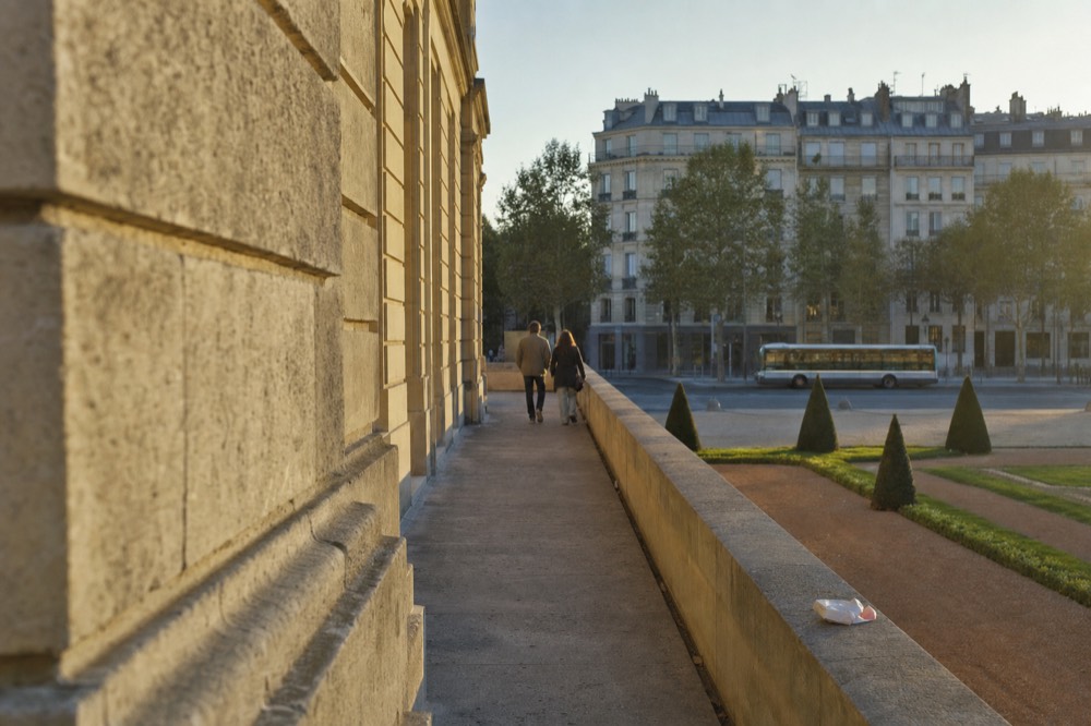 Un autobús urbano pasa por un amplio bulevar de París, más allá de una terraza de piedra soleada, jardines recortados, peatones lejanos y edificios Haussmann.