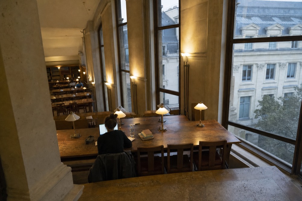 A reader sits with a laptop at a long wooden table in a tall-windowed library overlooking Parisian stone buildings.