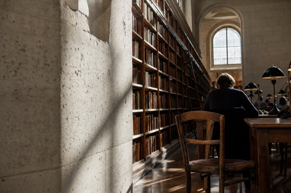 Sunlight falls across limestone walls, tall wooden bookshelves and desks in a historic reading room with a reader seen from behind.