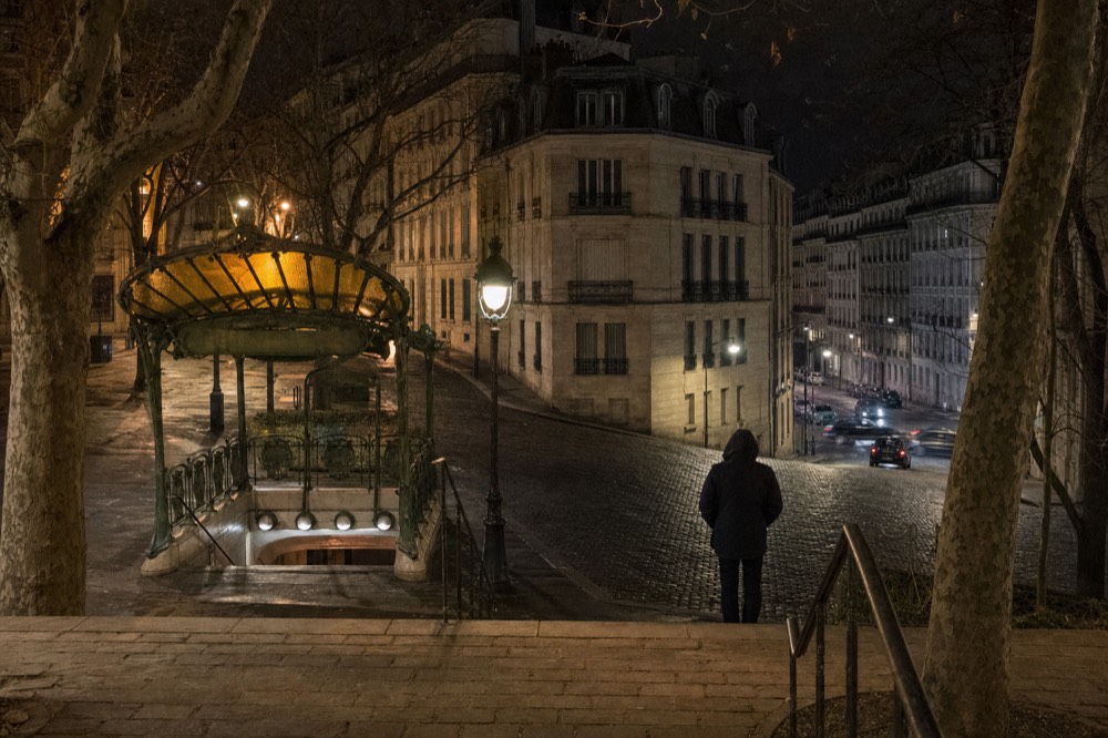 Amber-lit Art Nouveau metro entrance beside plane trees on a cobbled Paris street at night, with a hooded person seen from behind.