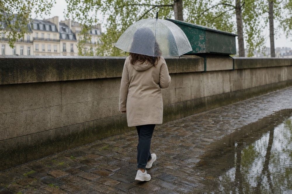 A person walks under a clear umbrella along a wet cobbled Seine quay beside a stone wall and green book boxes.