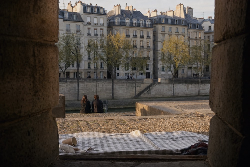 A picnic blanket and fallen paper cup lie on a gravel quay framed by stone walls, with Paris rooftops and riverside buildings beyond.