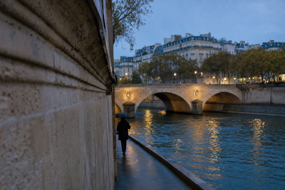 Rainy Seine riverbank at dusk with a stone bridge, lit reflections on the water, and a lone umbrella walker beside a limestone wall.