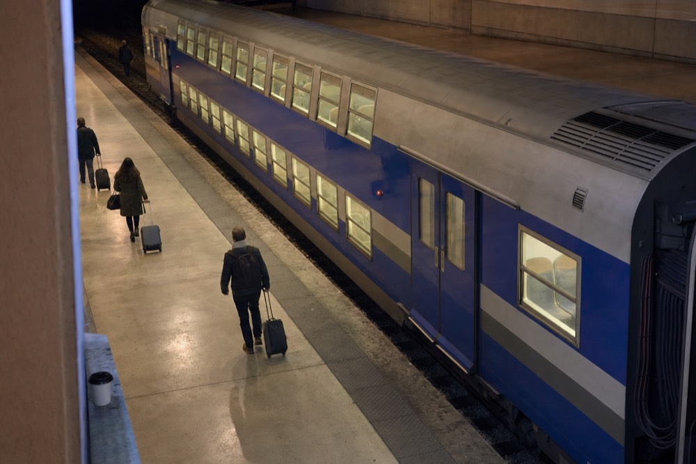 Travelers with luggage walk beside a blue double-deck train on a quiet concrete station platform.