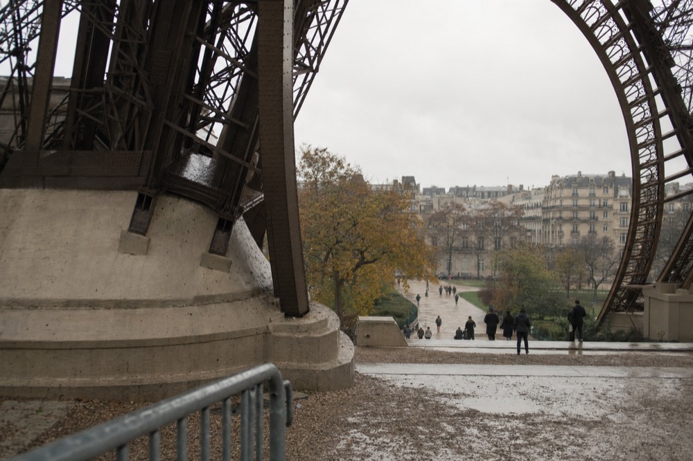 Wet paving and a barrier sit beneath the Eiffel Tower legs, with autumn trees, distant people, and Parisian buildings beyond.