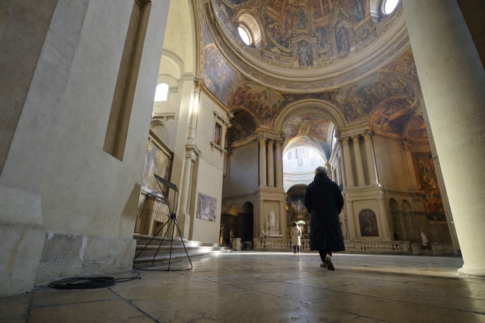 A low view inside a stone nave shows columns, a frescoed dome, a floor cable coil, and two distant visitors.