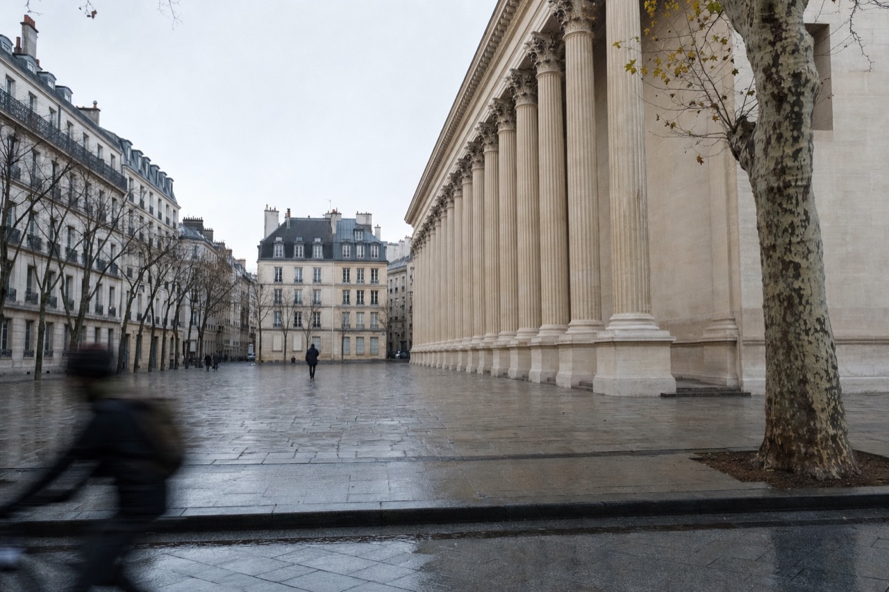 Wet paving reflects the Pantheon colonnade beside a plane tree, with Paris apartment blocks and a blurred cyclist in the square.