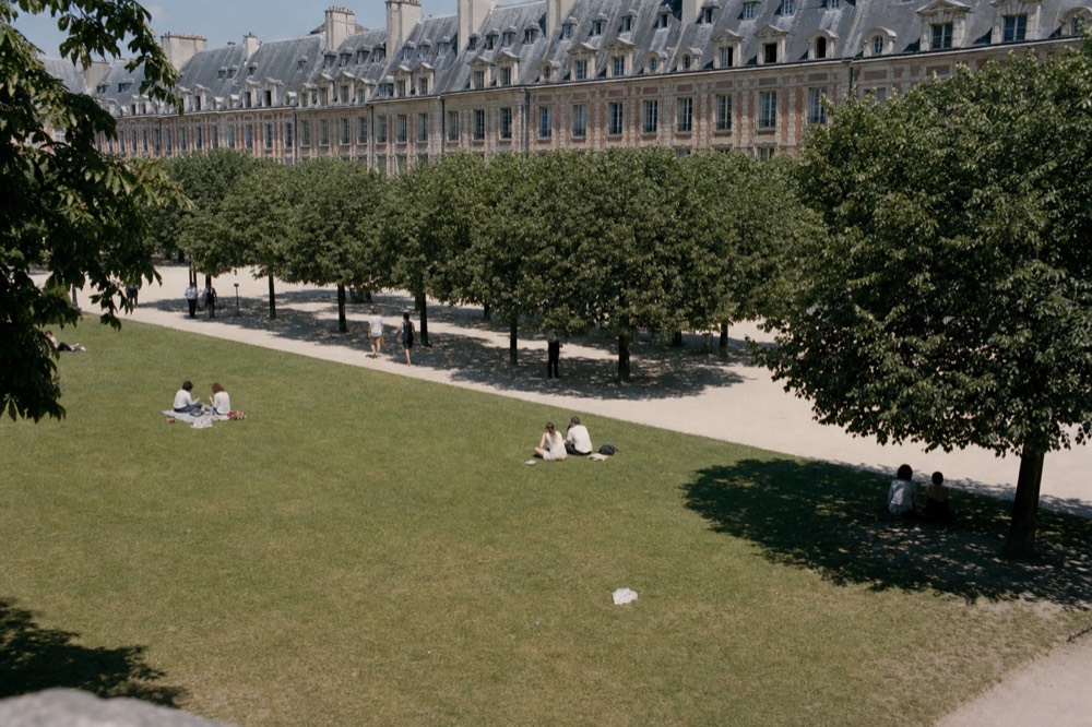 La gente sentada en un césped soleado de un parque de París, bajo árboles podados y edificios con arcadas de ladrillo y techos de pizarra con mansardas.