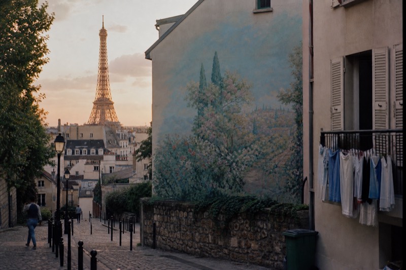 La Torre Eiffel iluminada por la luz cálida del atardecer sobre los tejados de zinc de París, vista gratis desde un callejón con escaleras junto a un mural pintado en una pared a dos aguas.