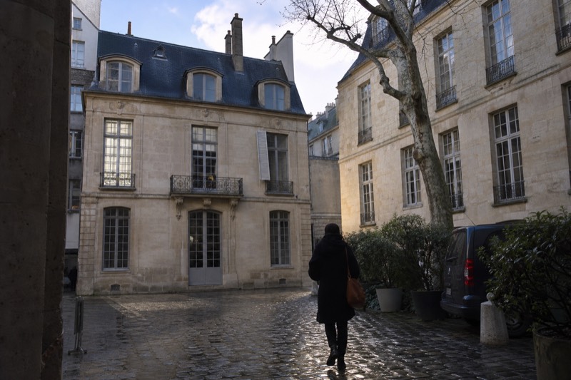 Un patio empedrado y tranquilo detrás de una casa señorial del siglo XVII en Le Marais, con paredes de piedra caliza clara, ventanas francesas altas y un plátano de sombra deshojado en la esquina.