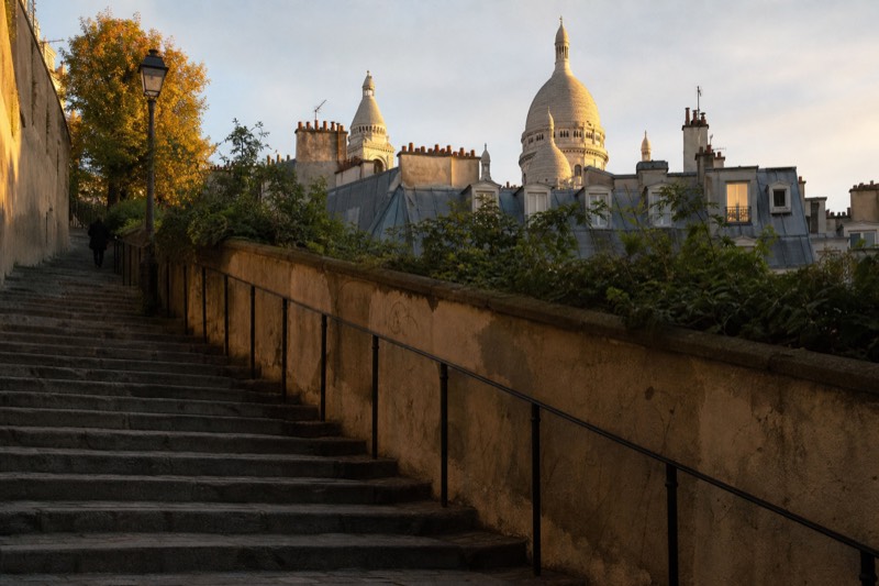 Escalones de piedra gastados que suben la colina de Montmartre bajo la luz tenue de una tarde de septiembre, con techos de zinc que descienden y la cúpula del Sacré-Cœur captando los últimos rayos de sol.