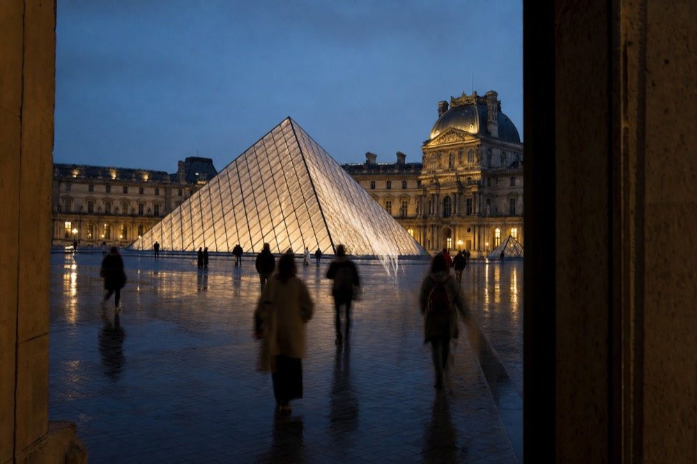 The Louvre pyramid glows at dusk in a wet courtyard with blurred visitors walking between dark stone opening edges.