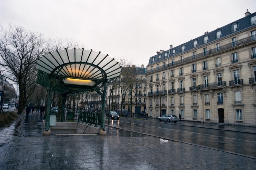 Rainy Paris boulevard with a green cast-iron metro entrance canopy, wet pavement, bare trees and limestone apartment facades.