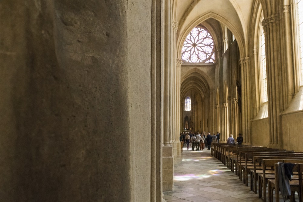 Gothic church nave with stone arches, pews, distant visitors, a rose window, and colored light on the floor.