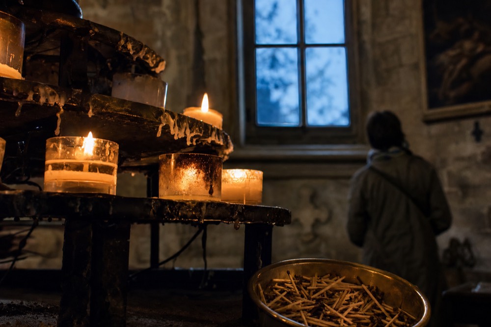 Lit votive candles on a dark iron stand beside a brass bowl of spent matches in a dim stone church chapel.