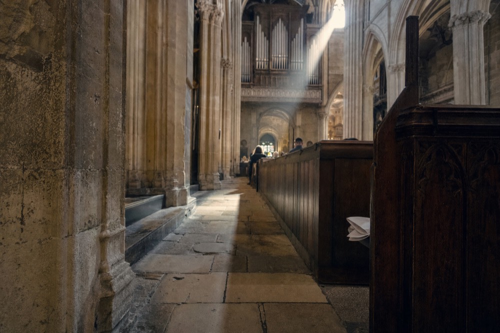 Organ pipes rise above a Gothic stone nave with a sunbeam crossing pews and a folded paper on a wooden pew end.