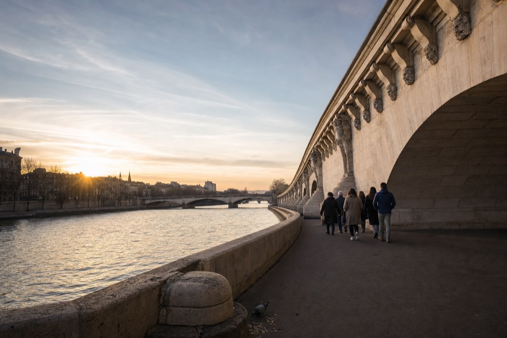 A small group walks beside the Seine under a stone Paris bridge at sunset, with arches, carved faces, water and a pigeon nearby.