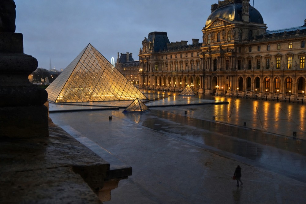 The Louvre glass pyramid glows at dusk in a wet palace courtyard with reflected lights and a lone distant figure.