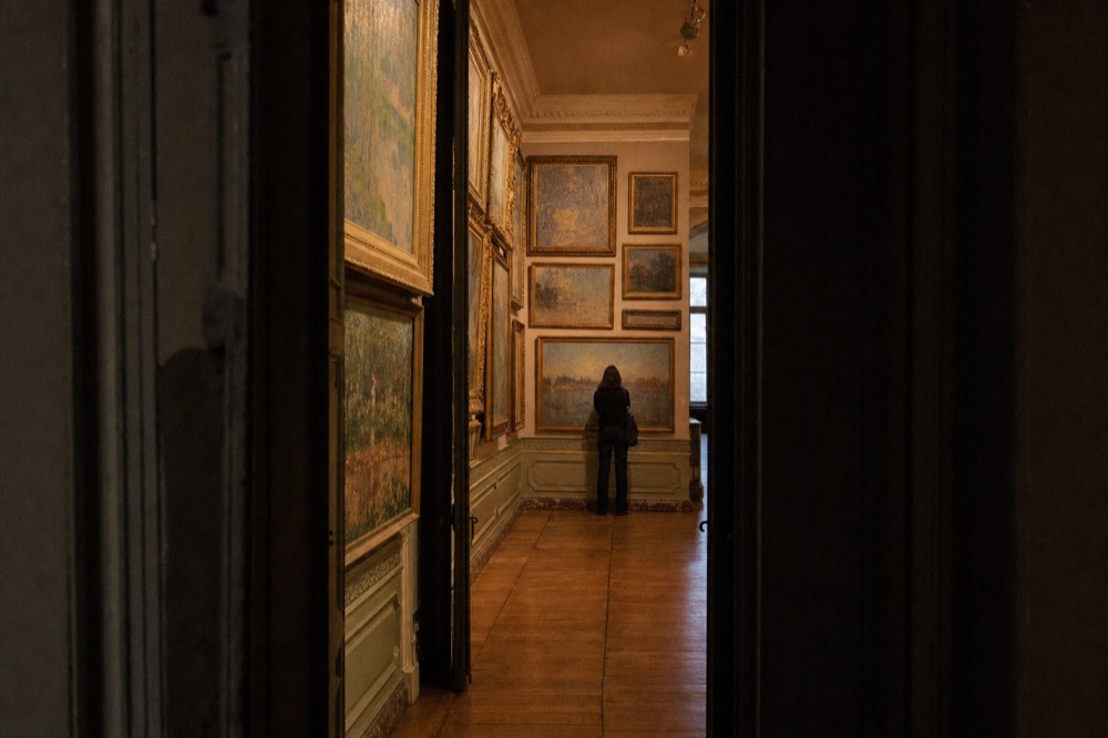 A dim museum gallery with ornate walls, many framed paintings, parquet flooring, and one visitor standing with their back turned.
