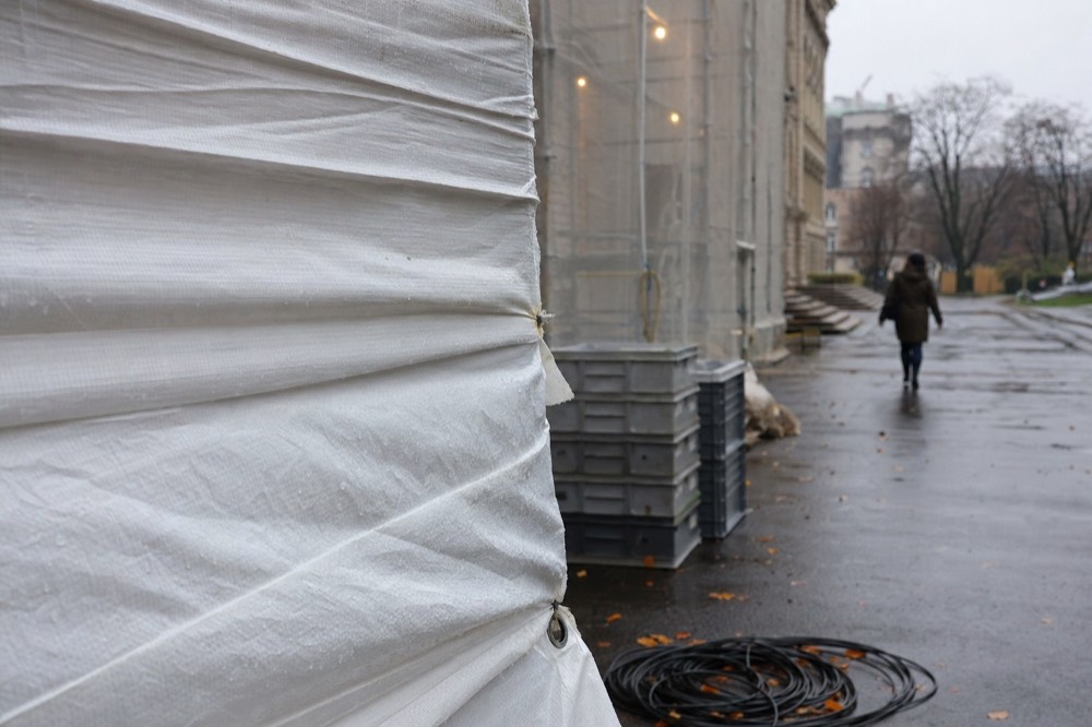 White tarpaulin covers scaffolding beside a limestone building, with plastic crates and a coil of cable on wet pavement.