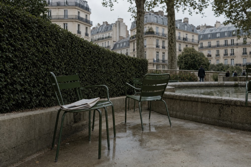 Green metal chairs sit beside a stone fountain basin and clipped hedge with Paris apartment buildings behind.