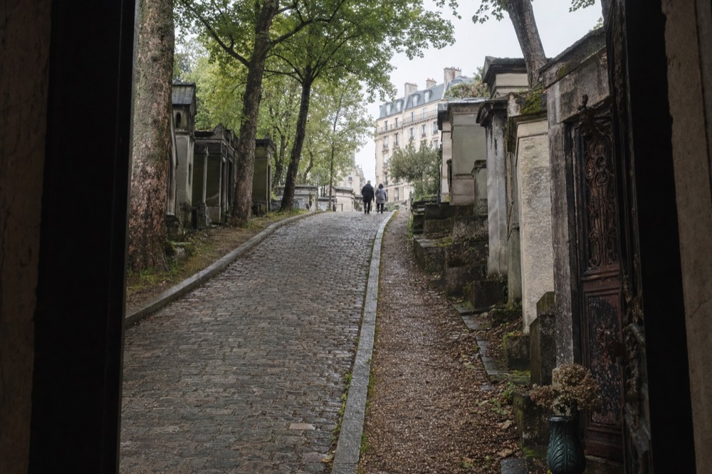 A cobbled cemetery lane climbs between old stone tombs and tall trees, with an iron door and distant people under a grey sky.