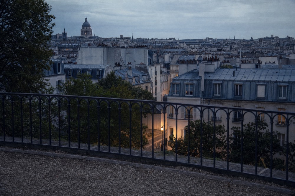 A dusk view over Paris rooftops and trees from a gravel terrace behind a dark iron railing, with a lit streetlamp below.