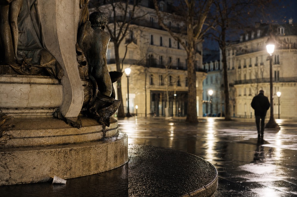 Bronze fountain figures stand beside wet paving, lamplight, bare trees and Paris stone buildings at night.