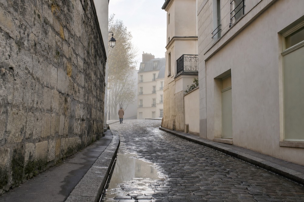 A narrow wet cobblestone lane curves between stone and cream Paris facades in morning fog with one distant pedestrian.