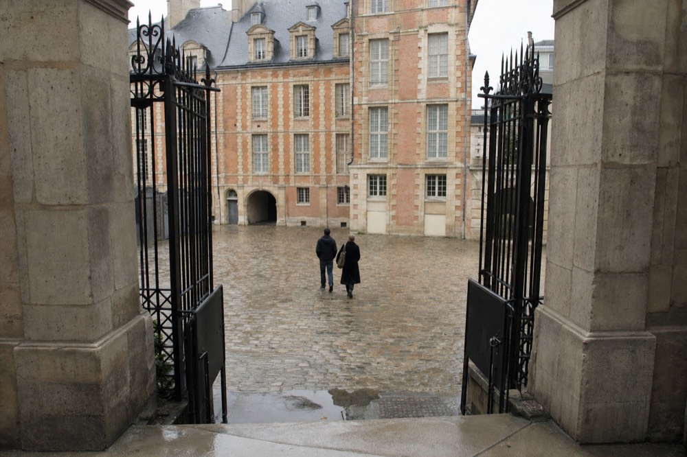 Open wrought-iron gates frame a cobbled courtyard with rose brick and pale stone facades, with two people walking away.