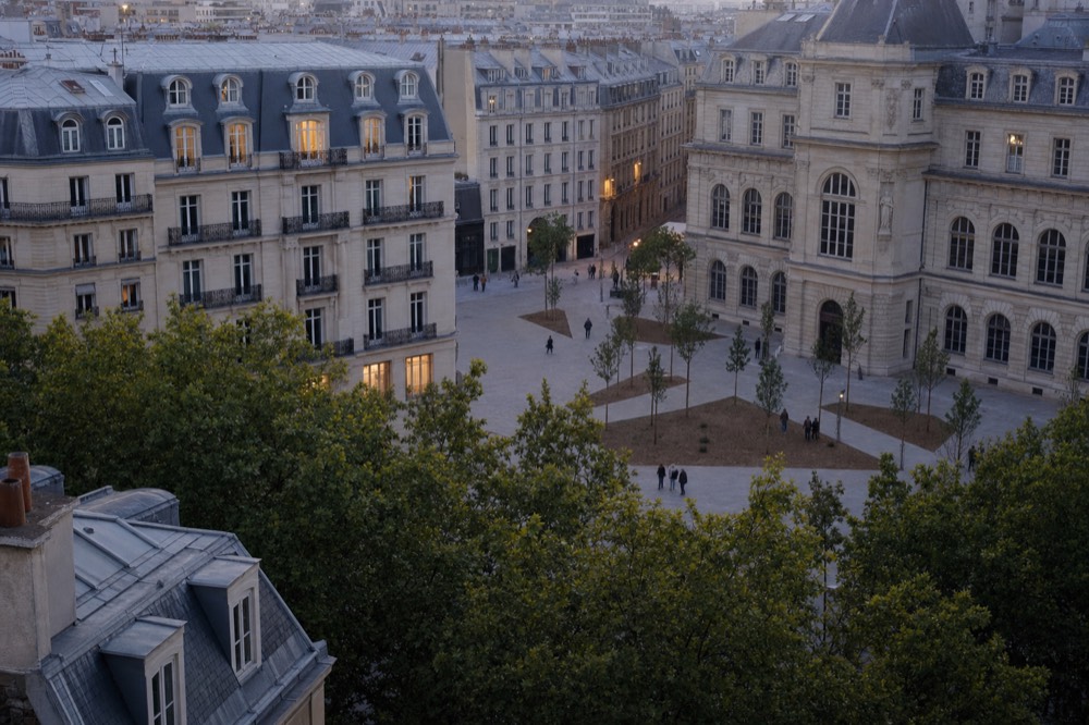 High view over Paris rooftops and trees toward a stone civic facade, young trees, and small distant pedestrians in a paved square at dusk.