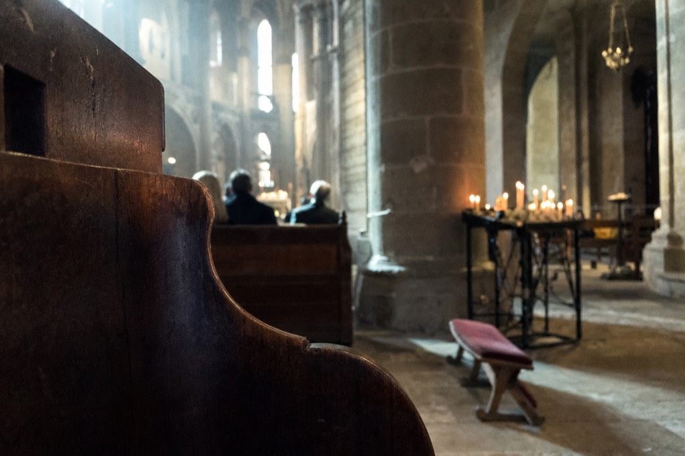 A wooden pew faces stone church pillars, distant seated people, a candle stand, and a kneeler in soft window light.