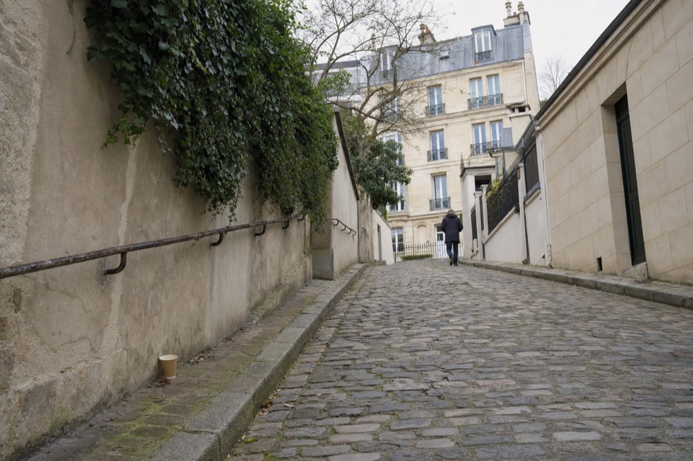 A steep cobbled Paris lane climbs between ivy-covered walls toward cream apartment buildings, with a distant person walking uphill.