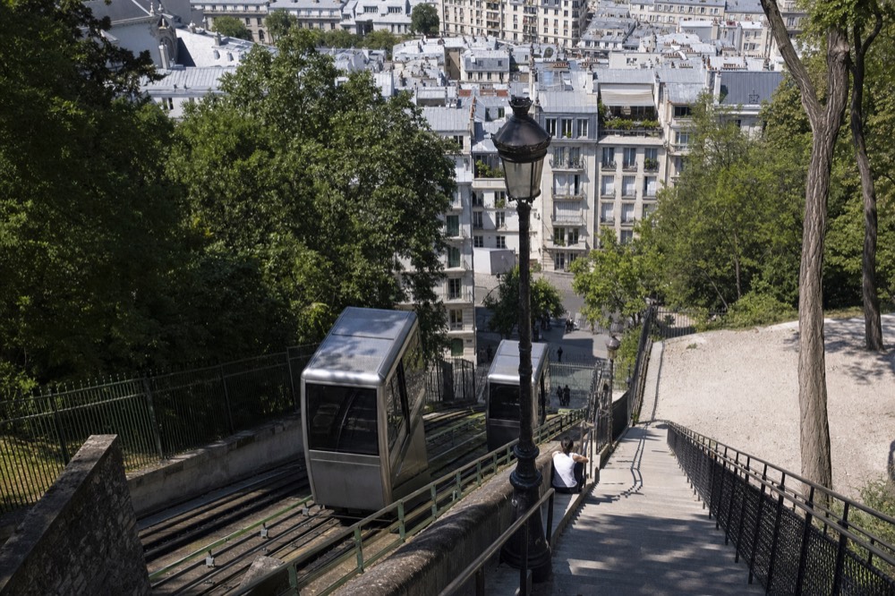 Montmartre funicular cars run beside a steep stone staircase with Paris rooftops and trees beyond.