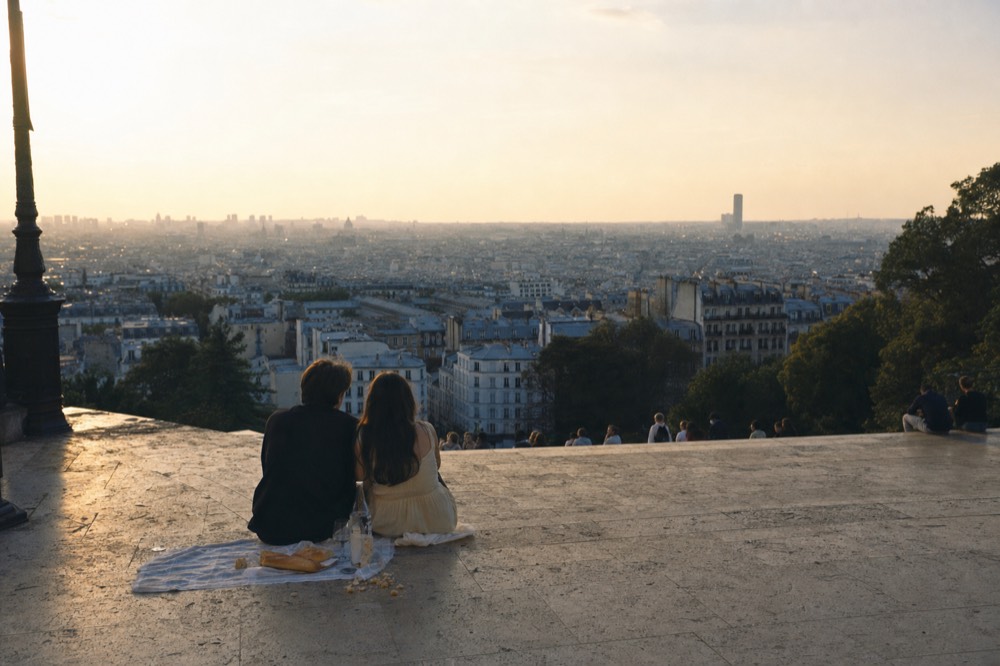 Two people sit with a picnic blanket and baguette on broad stone steps overlooking the hazy Paris skyline at sunset.