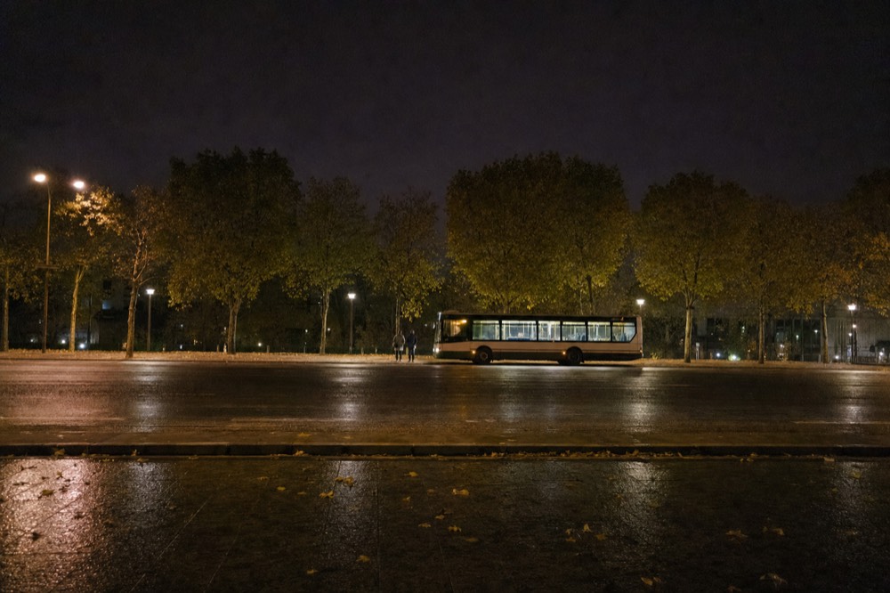 A long bus waits across a wet boulevard at night under streetlights and plane trees, with two distant pedestrians nearby.