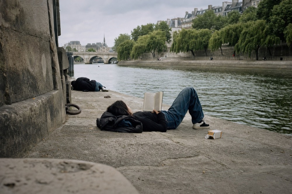 A person lies reading on a stone Seine quay, with another figure farther along the river and Paris buildings beyond.